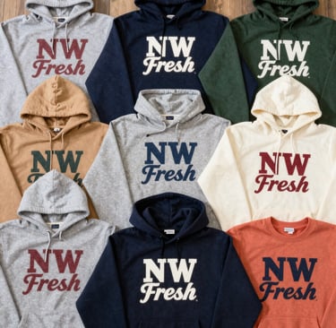 A flat lay of hoodies in earthy tones spread over a wooden table with pinecones and fallen leaves.
