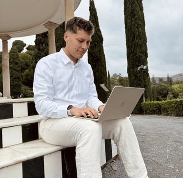 A man in a white shirt and linen pants working on a silver laptop outdoors in a park.