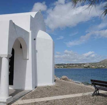 White Greek Orthodox church by the sea with a wooden bench overlooking the Aegean coast.
