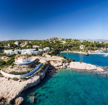 Aerial view of a coastal Greek village with white houses, turquoise sea, and rocky cliffs under a clear blue sky.