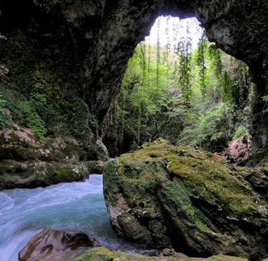 A turquoise river flows through a lush, mossy limestone cave opening in a forest landscape.