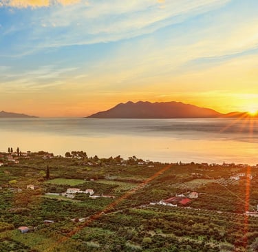 Golden sunset over a coastal village and lush green groves with mountains and calm sea in the background.