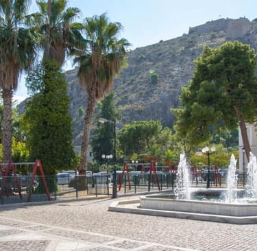 A sunny public square with palm trees and a tiered water fountain below a hilltop fortress.