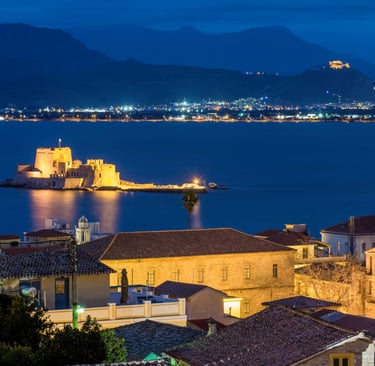 Illuminated Bourtzi Castle in Nafplio bay at night with city lights and mountains.