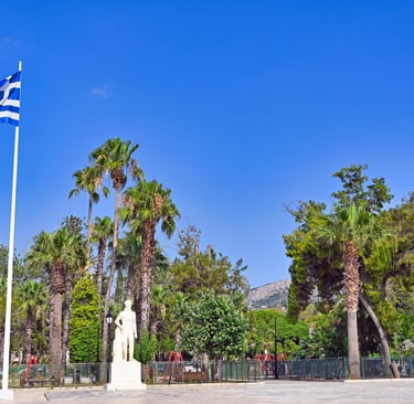 A Greek flag waving in a sunny park with palm trees and a white marble statue in Nafplio.