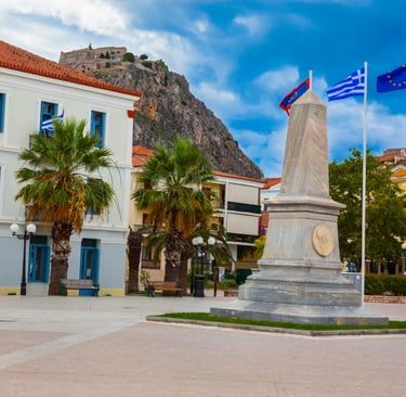A marble monument in Nafplio square with palm trees and Palamidi Fortress on the hill.