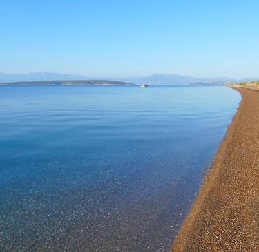 Calm clear blue sea meeting a pebble beach shoreline with distant mountains under a bright sky.