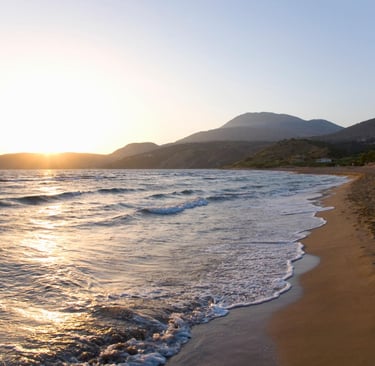 Golden sunset over a sandy beach with ocean waves and mountain landscape in the background.