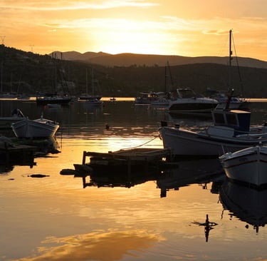 Small fishing boats anchored in a calm bay at sunset with mountain silhouettes in the background.