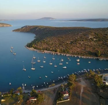 Aerial view of sailboats anchored in a calm blue Mediterranean bay near a lush coastline.