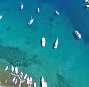 Aerial view of sailboats and motorboats anchored in a crystal clear turquoise bay along a tropical coastline.