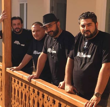 Four men in matching black Bohème t-shirts posing on a wooden balcony railing outdoors.