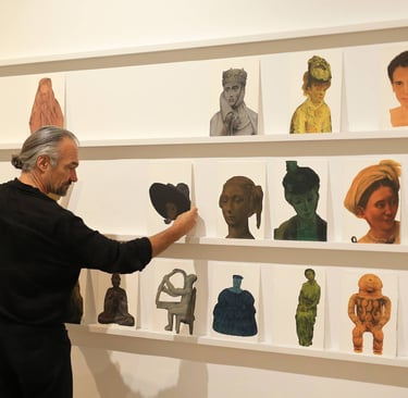 An artist arranges a diverse collection of portrait paintings on white gallery shelves for an exhibition.