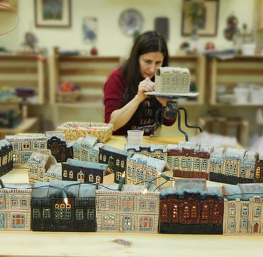 A ceramic artist handcrafting miniature clay houses and glowing ceramic city buildings in a pottery studio.