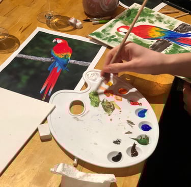 A person using a paint palette and brush to create a colorful scarlet macaw parrot acrylic painting.