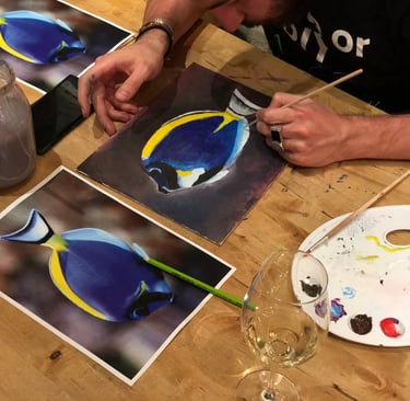 An artist using a paintbrush to create a realistic oil painting of a blue tang fish on a wooden table.