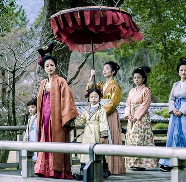 Women in traditional Japanese hanfu and kimono walk on a wooden bridge under a red ceremonial umbrella.