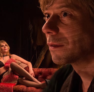 A blond man stares intently while a woman in lingerie poses on a red leather sofa in a dimly lit room.