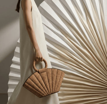Woman in white dress carrying a beige cork fan-shaped handbag against a pleated background.