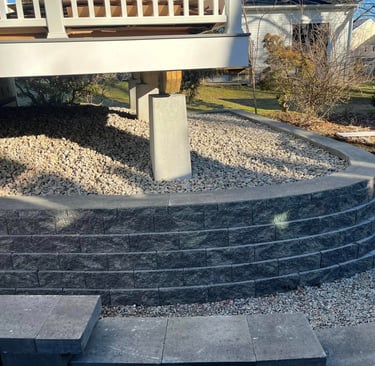 Curved grey block retaining wall supporting a house deck with gravel drainage and concrete piers.