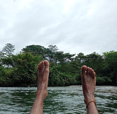 Bare feet relaxing over a flowing river with a background of lush tropical green trees and a cloudy sky.