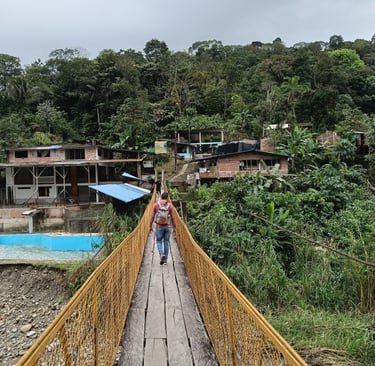A hiker crosses a narrow wooden suspension bridge towards a hillside village in the tropical rainforest.