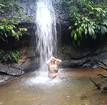 A woman bathing under a natural tropical waterfall surrounded by lush green jungle foliage.