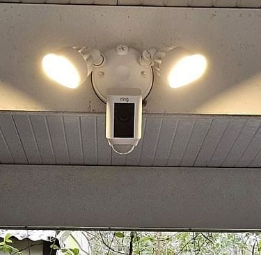 Ring floodlight security camera with dual LED lights installed on a white soffit for home surveillance.