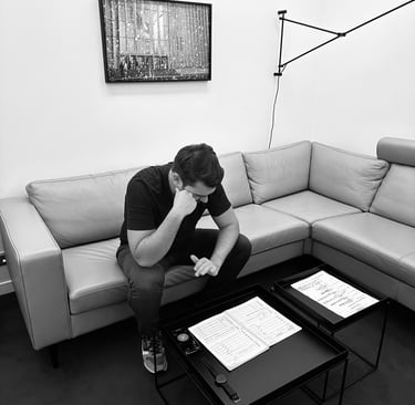 A man sits on a grey sectional sofa reviewing music sheets on a minimalist black coffee table.