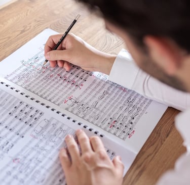 A composer writing and editing a musical score with a pencil on a wooden desk.