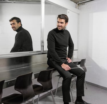 A male conductor in a black turtleneck sitting in a dressing room holding an orchestra baton.