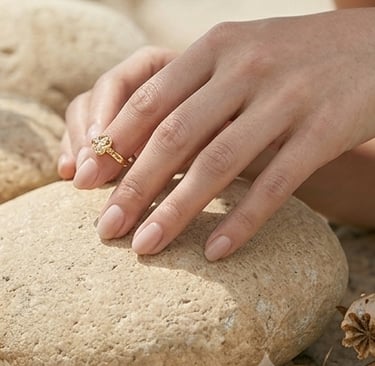 Mano con manicura en tono nude y un anillo de trébol dorado, apoyada sobre una piedra natural lisa.