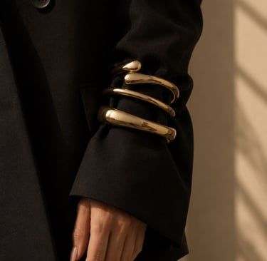 Gold statement bracelets worn over an oversized black blazer sleeve in soft natural light.