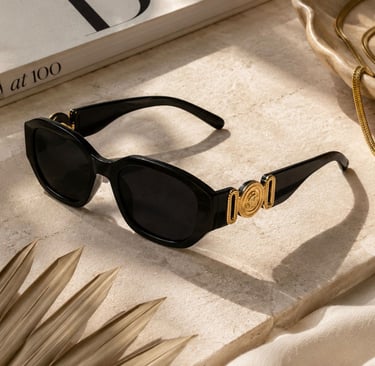 Black luxe sunglasses with gold detailing displayed on a beige stone surface