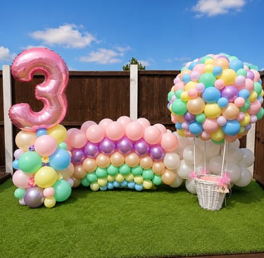 Pastel rainbow balloon arch and hot air balloon basket for a third birthday party decoration outdoors.