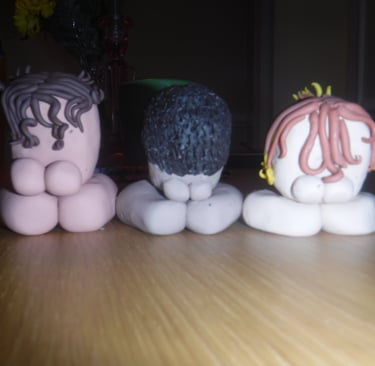 Three small clay figures with different hair textures sit on a wooden table in a dimly lit room.