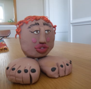 Handmade clay sculpture of a person's head with orange hair sitting on two large feet.