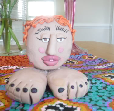 Handmade clay head sculpture with orange hair and feet base on a colorful patterned tablecloth.