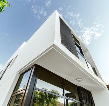 Low angle view of a modern white minimalist house with large glass windows and black shutters against a blue sky.