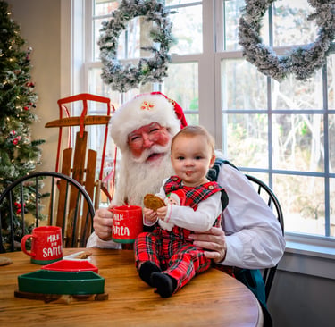 Santa Claus sitting at a wooden table with a baby in plaid overalls holding Christmas cookies and milk mugs.