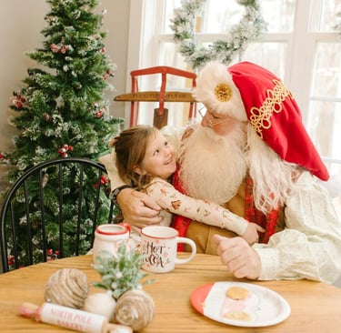 Smiling young girl hugging Santa Claus at a holiday table with cookies and a Christmas tree.