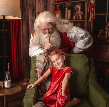 Santa Claus making a quiet gesture behind a smiling young girl sitting in a green armchair.