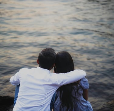 Couple sitting by the water with arms around each other during a romantic sunset date.