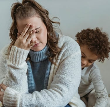 A stressed mother holding her head in frustration while her young child plays behind her at home.