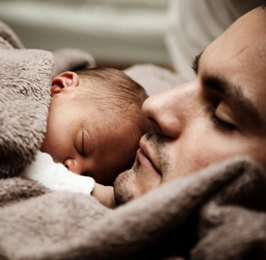 A newborn baby sleeping peacefully against their father's face under a soft brown blanket.