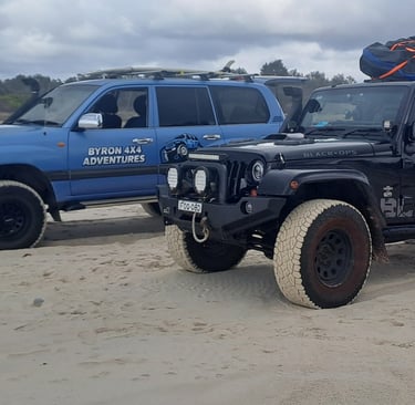 Black Jeep Wrangler and blue Toyota Land Cruiser 4x4 vehicles driving on a sandy beach for off-road adventures.