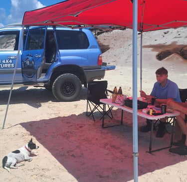 A blue Byron 4x4 Adventures truck parked on a beach with people dining under a red canopy tent.