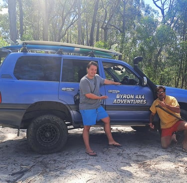 Two men posing next to a blue Byron 4x4 Adventures Toyota Land Cruiser with surfboards in a forest.