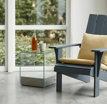 Modern grey Adirondack chair with yellow pillows and a glass side table in a bright sunroom.