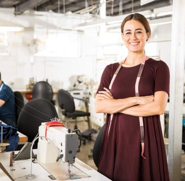 Un diseñador de moda sonriente con una cinta métrica, de pie en una fábrica de ropa con máquinas.